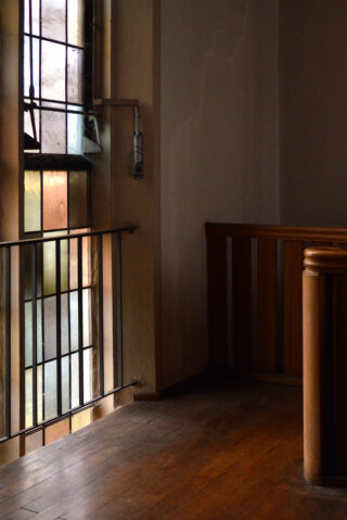 Ein sonnenbeschienenes Buntglasfenster wirft farbiges Licht auf einen Holzboden neben einem Holzgeländer in einem ruhigen, leeren Innenraum.