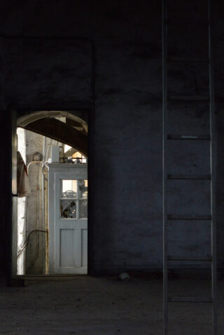 An einer dunklen Wand steht eine Metallleiter, während eine alte Holztür offen steht und den Blick auf einen sonnenbeschienenen Bereich und eine weitere geschlossene Tür dahinter freigibt.