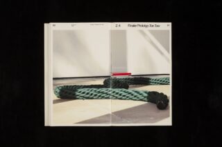 In einem offenen Buch ist das Foto eines dicken grünen Seils zu sehen, das auf einem hellen Boden in der Nähe einer weißen Wand und eines kleinen roten Objekts aufgerollt ist.