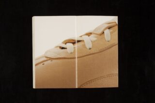 Auf den aufgeschlagenen Seiten eines Buches ist auf schwarzem Hintergrund die Nahaufnahme eines hellbraunen Leder-Sneakers mit weißen Schnürsenkeln gedruckt.