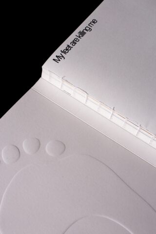 Ein offenes Buch mit einem geprägten Fußabdruck auf einer Seite und dem Text „Meine Füße bringen mich um“ in der Nähe des Buchrückens.