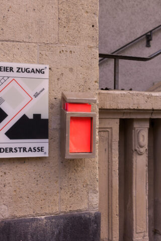 An einer Steinmauer ist neben einer Karte und einem Treppengeländer ein rechteckiges, beleuchtetes rotes Schild angebracht.