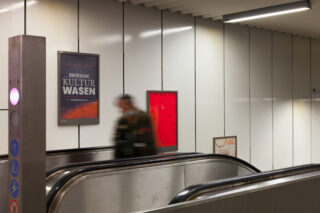 Eine Person in Bewegung steht auf einer Rolltreppe in einer U-Bahn-Station mit weiß getäfelten Wänden, einem Plakat und einem roten Notfallkasten.