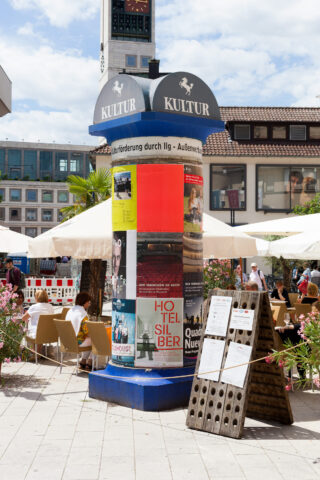 Eine zylindrische Litfaßsäule mit Plakaten steht in einem Café-Außenbereich, umgeben von Tischen, Stühlen, Sonnenschirmen und umliegenden Gebäuden.
