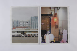 In einer aufgeschlagenen Zeitschrift sind zwei Fotos zu sehen: eines von einem Parkplatz mit Autos und hohen Gebäuden und eines von einer engen Stadtgasse mit einer roten Laterne und verschiedenen Gegenständen.