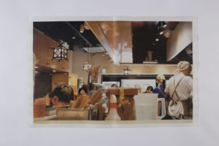 Eine geschäftige Restaurantküche mit arbeitendem Personal, von denen einige Schürzen und Kopftücher tragen, die hinter einer Theke Essen zubereiten, wobei Utensilien und Vorräte sichtbar sind.