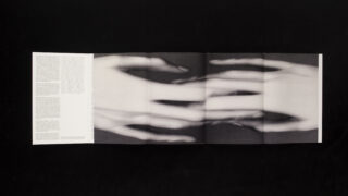 Auf einer ausklappbaren Broschüre ist ein verschwommenes Schwarzweißbild mit überlappenden Händen zu sehen. Der Text ist auf der äußersten linken Seite auf schwarzem Hintergrund abgedruckt.