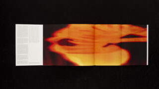 Ein dreifach gefaltetes Heft auf einer schwarzen Oberfläche zeigt verschwommene orange und gelbe abstrakte Kunst auf drei Tafeln, wobei auf der äußersten linken Tafel Text aufgedruckt ist.