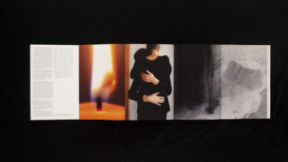 Ein gefaltetes Heft auf einer schwarzen Oberfläche zeigt Text, eine verschwommene Flamme, zwei sich umarmende Menschen und ein abstraktes Graustufenbild auf vier Tafeln.