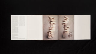 Ein offenes Heft zeigt Text auf der linken Seite und zwei mittlere Seiten mit unscharfen, vertikalen Stapeln von Blöcken auf einem neutralen Hintergrund, mit einer leeren rechten Seite.