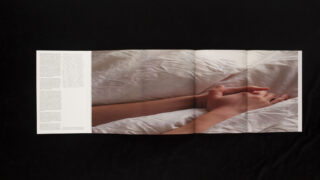 Eine aufgeschlagene Zeitschrift auf einer dunklen Oberfläche zeigt ein Foto von zwei gefalteten Händen auf weißem Bettzeug, mit Text auf der linken Seite.