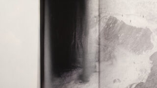 Ein zweiseitiges Schwarzweiß-Luftbild zeigt eine Küstenlinie mit dem Meer auf der linken Seite und einem Strand mit kleinen Figuren in der Ferne auf der rechten Seite.