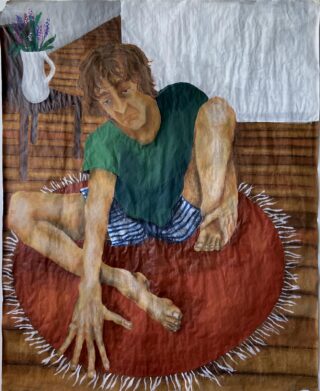 Eine Person mit hellbraunem Haar sitzt im Schneidersitz auf einem runden roten Teppich. Sie trägt ein grünes Hemd und gestreifte Shorts und steht neben einem Holztisch mit einer weißen Blumenvase.