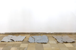 Drei große, unregelmäßig geformte graue Platten sind auf einem gefliesten Boden vor einer schlichten weißen Wand angeordnet.