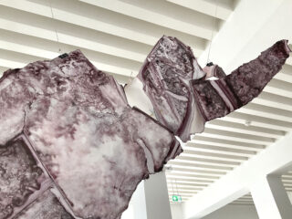 Eine große, abstrakte Skulptur in marmorierten Rosa- und Lilatönen hängt in einem modernen Galerieraum mit weißen Wänden von der Decke.