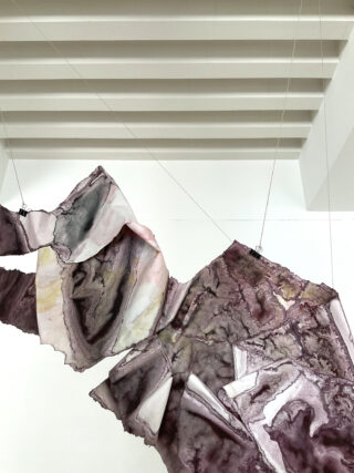 Große, abstrakte, stoffähnliche Paneele mit marmorierten Mustern in Lila, Rosa und Grau hängen an Drähten vor einer weißen Galeriewand und -decke.