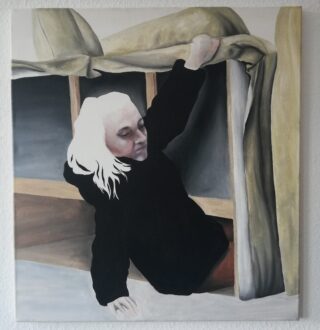 Eine Person mit hellem Haar und schwarzem Oberteil lehnt sich in einem sanft beleuchteten Raum unter einer hochgezogenen Decke oder einem Vorhang hervor und hält den Stoff über ihrem Kopf.