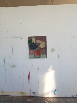 Ein kleines abstraktes Gemälde ist auf einer großen, weißen Wand mit sichtbaren Flecken, Markierungen und Farbtropfen zentriert. Der Boden ist unten teilweise sichtbar.