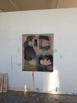 Ein abstraktes Gemälde mit überlappenden, gedämpften Formen und Figuren ist an einer weißen Wand in einem Kunstatelier angebracht, wobei Farbflecken an Wand und Boden sichtbar sind.