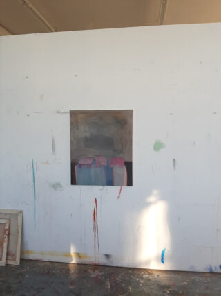 An einer weißen, mit Farbe bespritzten Wand hängt ein Gemälde; das Kunstwerk zeigt vier abstrakte, bunte Formen vor einem dunklen Hintergrund.