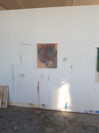 Ein einzelnes abstraktes Gemälde in Rot- und Grautönen hängt an einer mit Farbe bespritzten weißen Wand im Atelier eines Künstlers und wird vom Sonnenlicht in einen Schatten geworfen.