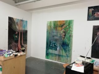 Kunstatelier mit unvollendeten Gemälden an weißen Wänden, einem Arbeitstisch mit Künstlerbedarf und verschiedenen im Raum verstreuten Materialien.