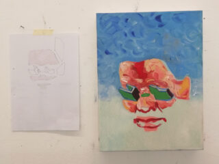 An einer Wand ist die Skizze eines abstrakten Gesichts neben einer bunt gemalten Version desselben Gesichts in Blau-, Orange- und Grüntönen aufgeklebt.