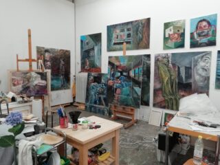 Ein Kunstatelier mit mehreren expressionistischen Gemälden auf Staffeleien und Wänden, umgeben von Kunstbedarf, Tischen und verschiedenen Materialien auf dem Boden.