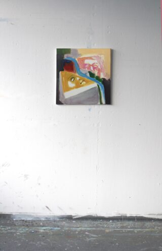 Ein kleines abstraktes Gemälde in kräftigen Farben hängt an einer weißen Wand über einem grauen, farbbespritzten Boden in einem Kunstatelier.