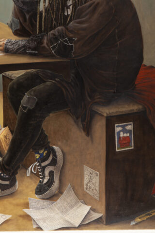 Eine Person mit langen Zöpfen und geflickter Kleidung sitzt an einem Schreibtisch; auf und um die Bank herum sind verstreute Papiere und kleine Zeichnungen zu sehen.