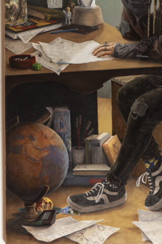 Eine Person sitzt an einem überladenen Schreibtisch, der mit Papieren, Büchern, Malutensilien und einem Globus übersät ist. Eine Hand schreibt, die andere ruht auf dem Schreibtisch. Nur die untere Gesichtshälfte und die Beine sind sichtbar.