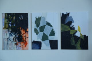 Drei abstrakte Gemälde werden nebeneinander auf einer weißen Wand ausgestellt, jedes weist geometrische Formen und kräftige, kontrastierende Farben auf.