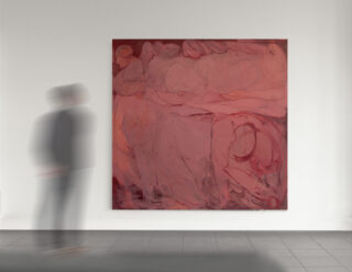 Eine in ihrer Bewegung verschwommene Person steht vor einem großen abstrakten Gemälde in Rot- und Rosatönen, das an einer weißen Galeriewand hängt.