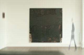 An einer weißen Galeriewand hängt ein großes, dunkles abstraktes Gemälde, auf der rechten Bildseite geht eine verschwommene Gestalt vorbei.