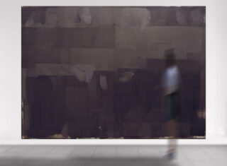 Eine Person geht an einem großen abstrakten Gemälde mit dunklen, rechteckigen Formen und gedämpften Tönen vorbei, das an einer weißen Wand in einer Galerie hängt.