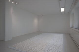 Ein minimalistischer, leerer weißer Raum mit einem strukturierten weißen Teppich in der Mitte des Bodens, Deckenleuchten darüber und Fenstern entlang der rechten Wand.