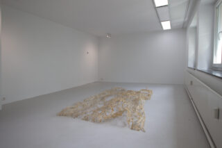 Ein minimalistischer Galerieraum mit weißen Wänden und Böden zeigt eine skulpturale Installation, die einer menschlichen Figur ähnelt und aus durchscheinendem, faserigem Material besteht, das auf dem Boden liegt.