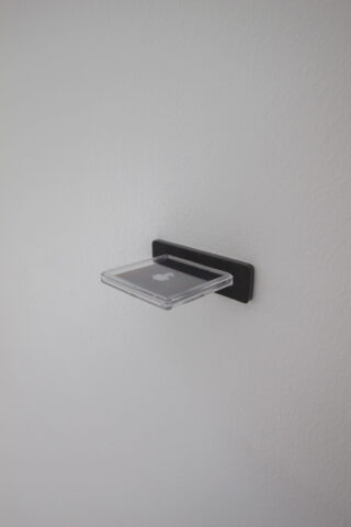 Ein kleines, transparentes rechteckiges Regal, das mit einer schwarzen Halterung an einer weißen Wand montiert wird.