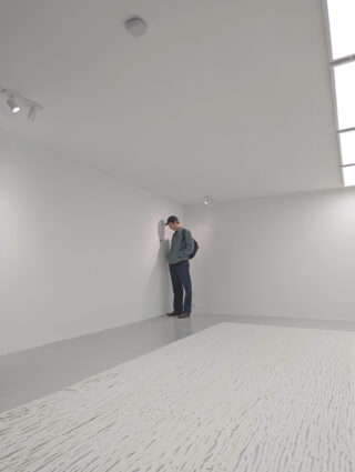 Eine Person steht allein in einem minimalistischen Raum mit weißen Wänden, blickt in die Ecke und lehnt ihren Kopf an die Wand.