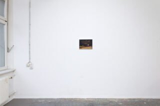 Ein kleines gerahmtes Bild hängt an einer weißen Wand in einem spärlich möblierten Raum mit Fenster, Heizkörper und freiliegenden Steckdosen und Leitungen.