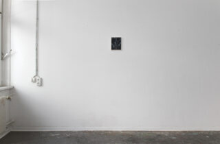 Ein kleines gerahmtes Bild hängt an einer großen, schlichten weißen Wand in einem weitgehend leeren Raum, in dem einige Rohre, Steckdosen und ein fleckiger Bodenbelag sichtbar sind.