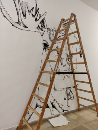 Eine Holzleiter steht vor einem teilweise fertiggestellten Wandbild mit schwarzen Linien auf einer weißen Wand. Es zeigt ein Tier und einen Menschen. Auf dem gefliesten Boden liegen eine Farbwanne und ein Pinsel.