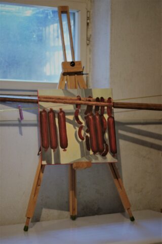 Ein Gemälde von an einer Stange hängenden Würstchen, ausgestellt auf einer Staffelei, mit einer echten Holzstange vor dem Gemälde, im Innenbereich in der Nähe eines Fensters.