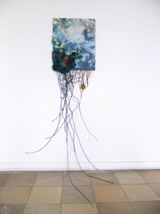 Ein Mixed-Media-Kunstwerk mit einer Leinwand mit abstrakten Bildern, künstlichen Pflanzen und nach unten verlaufenden farbigen Schnüren, befestigt an einer weißen Wand über einem gefliesten Boden.