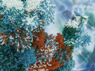 Nahaufnahme von strukturierten Garnpompons in Blau, Weiß, Blaugrün und Braun auf einem blau-weiß gemusterten Stoffhintergrund.