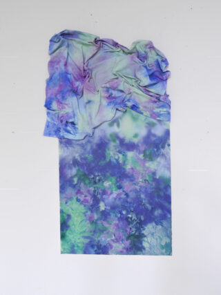 Ein Stück zerknitterter, pastellfarbener Stoff in Blau-, Lila- und Grüntönen liegt über einer bemalten Oberfläche mit ähnlichen Farben und abstrakten Mustern auf weißem Hintergrund.