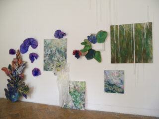 Eine Galeriewand zeigt abstrakte Kunstwerke mit botanischen Motiven, darunter Gemälde und dreidimensionale Blumenskulpturen, die asymmetrisch auf einer weißen Wand über einem Holzboden angeordnet sind.