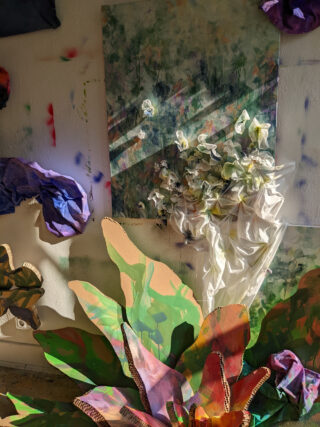 Ein Stillleben mit in Stoff gehüllten weißen Blumen, bunt bemalten Blättern und Sonnenlicht, das Schatten auf eine Wand mit abstrakten Farbspritzern wirft.