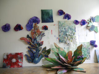 Eine weiße Wand zeigt farbenfrohe abstrakte Kunst und 3D-Papierskulpturen von Pflanzen und Blumen, während auf dem Boden und an der Wand bemalte Leinwände und Kartonstrukturen angeordnet sind.