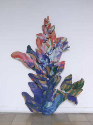 Vor einer weißen Wand steht eine große, abstrakte, bemalte Pappskulptur, die einer Pflanze mit geschichteten, bunten Blättern ähnelt.